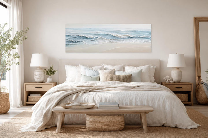 Wide coastal canvas art of soft blue ocean waves on a sandy beach above a neutral bedroom headboard.