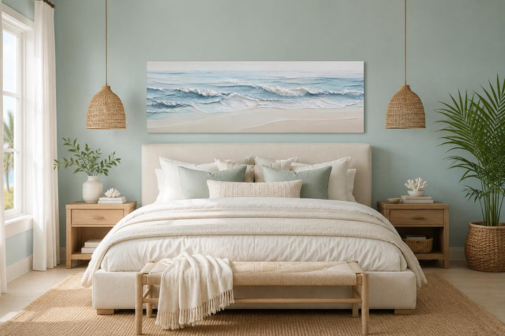 Calm ocean waves wall art in soft blue and cream tones hanging above a bed in a coastal bedroom.