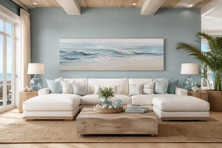 Wide horizontal canvas art of soft blue ocean waves on a sandy beach above a white sectional sofa.