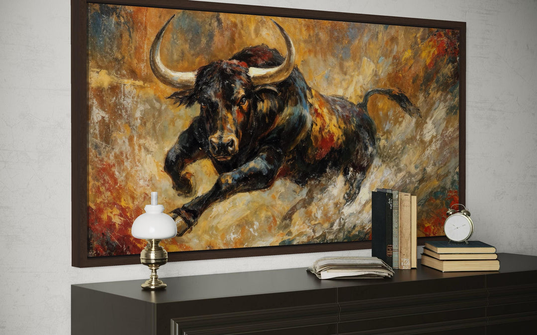 Charging bull canvas print in a dark frame above a dresser with books and a lamp.