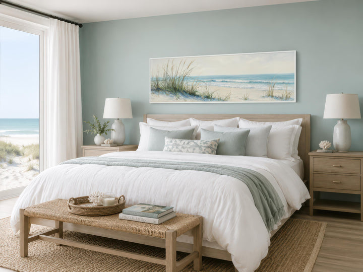 Wide horizontal canvas print of sandy beach dunes and ocean above a bed in a coastal bedroom.