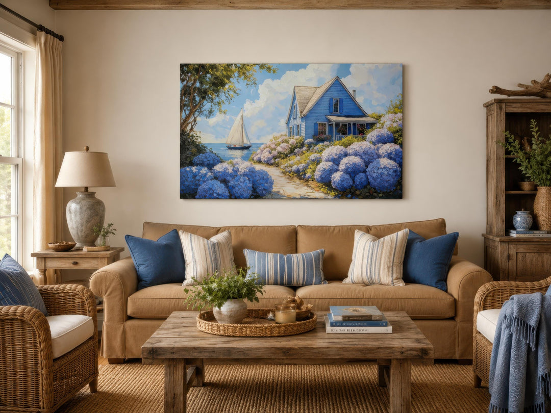 Coastal canvas print of a blue cottage and sailboat scene above a tan sofa in a living room.