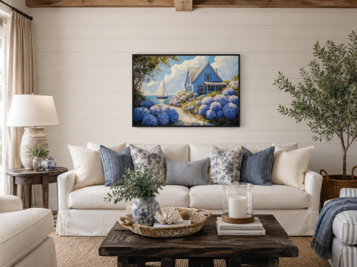 Coastal canvas print of a blue cottage and sailboat scene above a white sofa with blue pillows.
