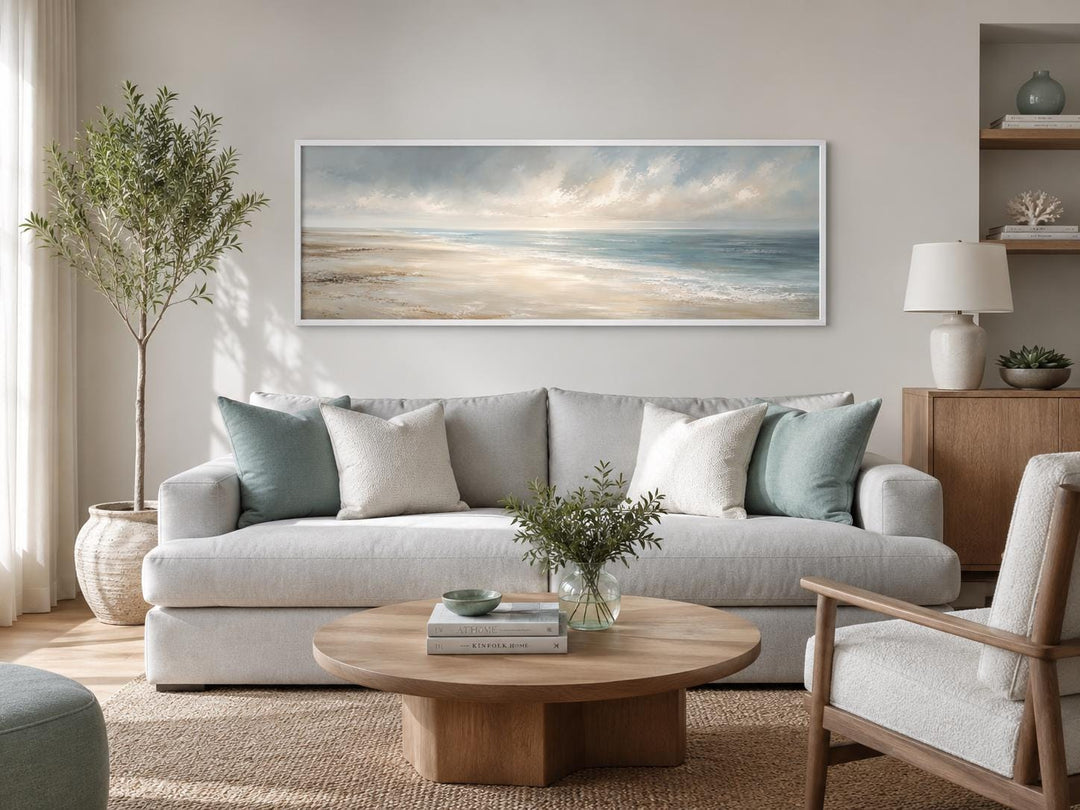 Long horizontal coastal wall art of a serene ocean seascape with soft clouds above a grey sofa.