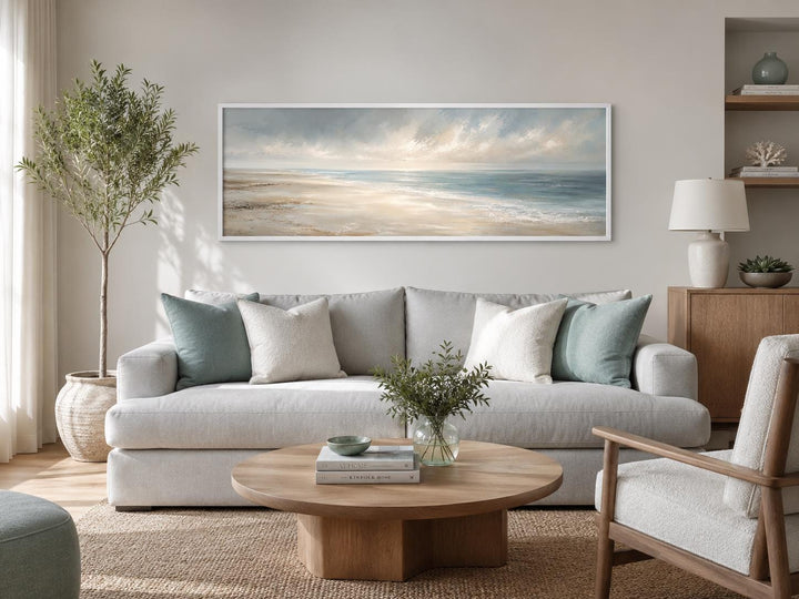 Long horizontal coastal wall art of a serene ocean seascape with soft clouds above a grey sofa.