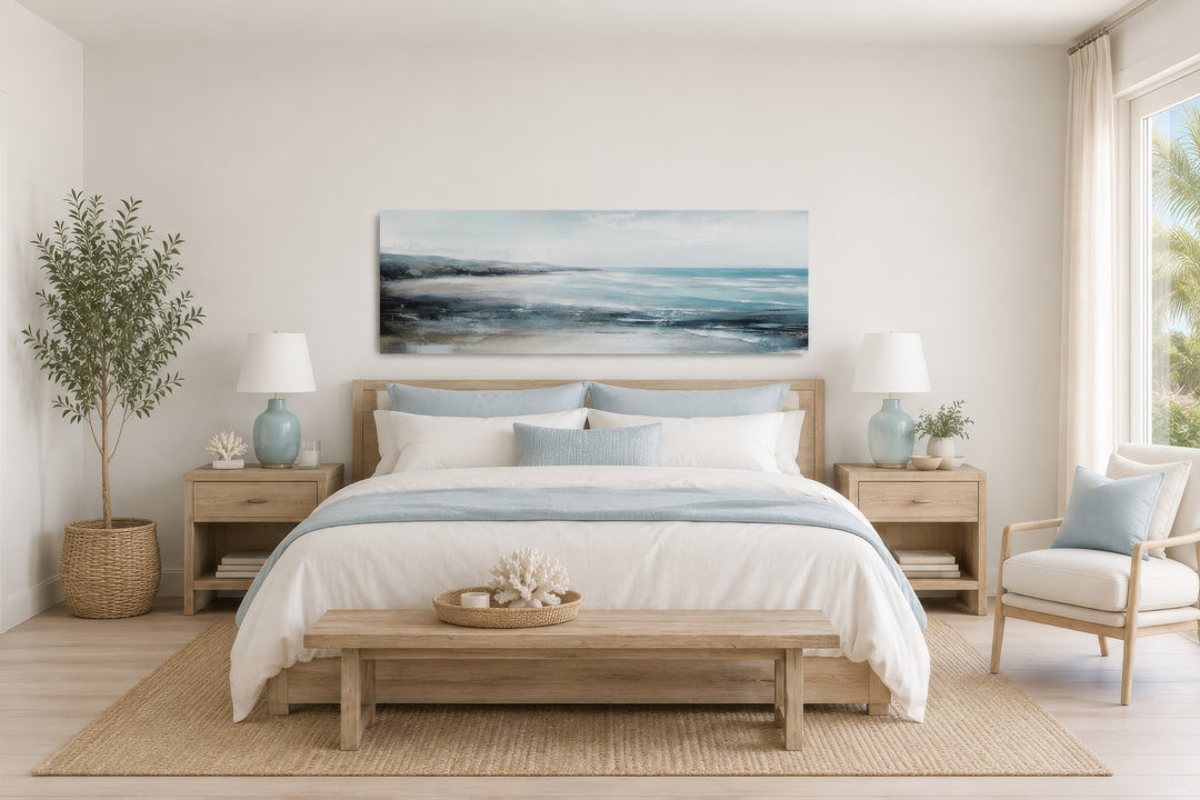 Long narrow coastal wall art of a serene ocean beach scene hanging over a bed in a neutral bedroom.