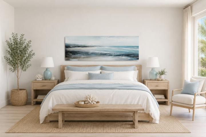 Long narrow coastal wall art of a serene ocean beach scene hanging over a bed in a neutral bedroom.