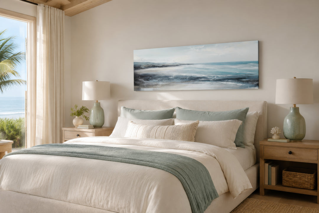 Long narrow coastal wall art of an ocean beach scene hanging over a bed in a neutral bedroom.