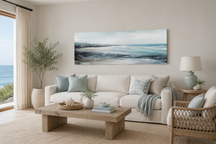 Long narrow coastal wall art of a serene ocean beach scene hanging over a white sofa.