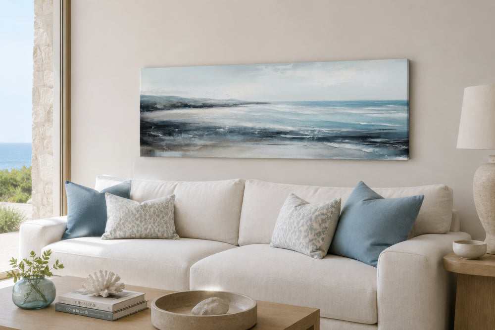 Long narrow coastal wall art featuring a serene ocean beach scene hanging above a white sofa.