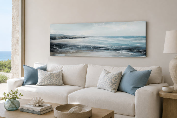 Long narrow coastal wall art featuring a serene ocean beach scene hanging above a white sofa.