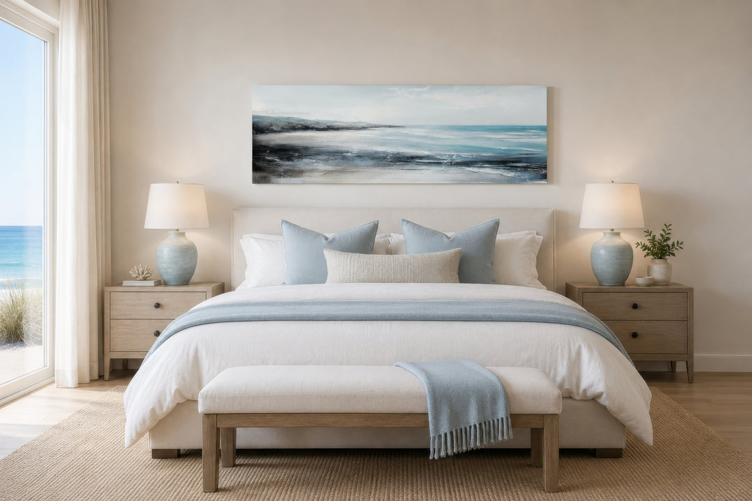 Long narrow coastal wall art of a blue ocean beach scene hanging over a bed in a neutral bedroom.