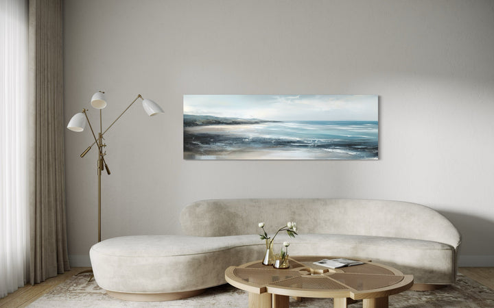 Long narrow coastal canvas wall art of a serene ocean beach scene above a modern curved sofa.