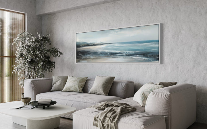Long narrow coastal wall art of a blue ocean beach scene hanging over a modern grey sectional sofa.