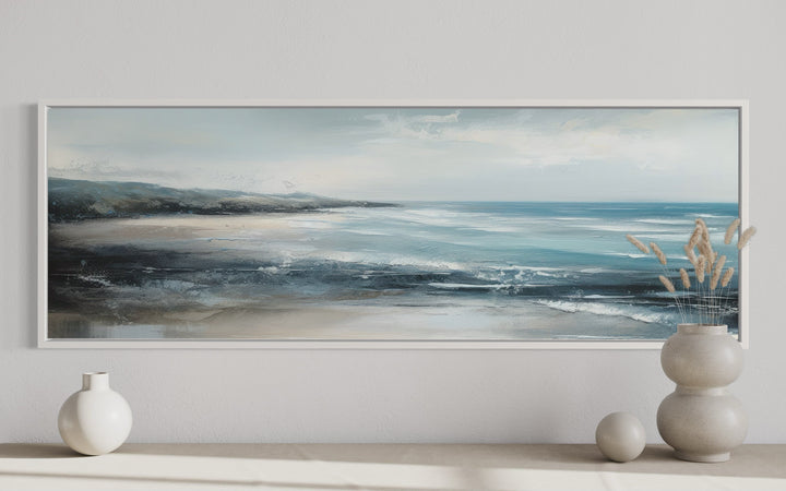 Long narrow coastal canvas art of a blue ocean and sandy beach in a white frame over a console.