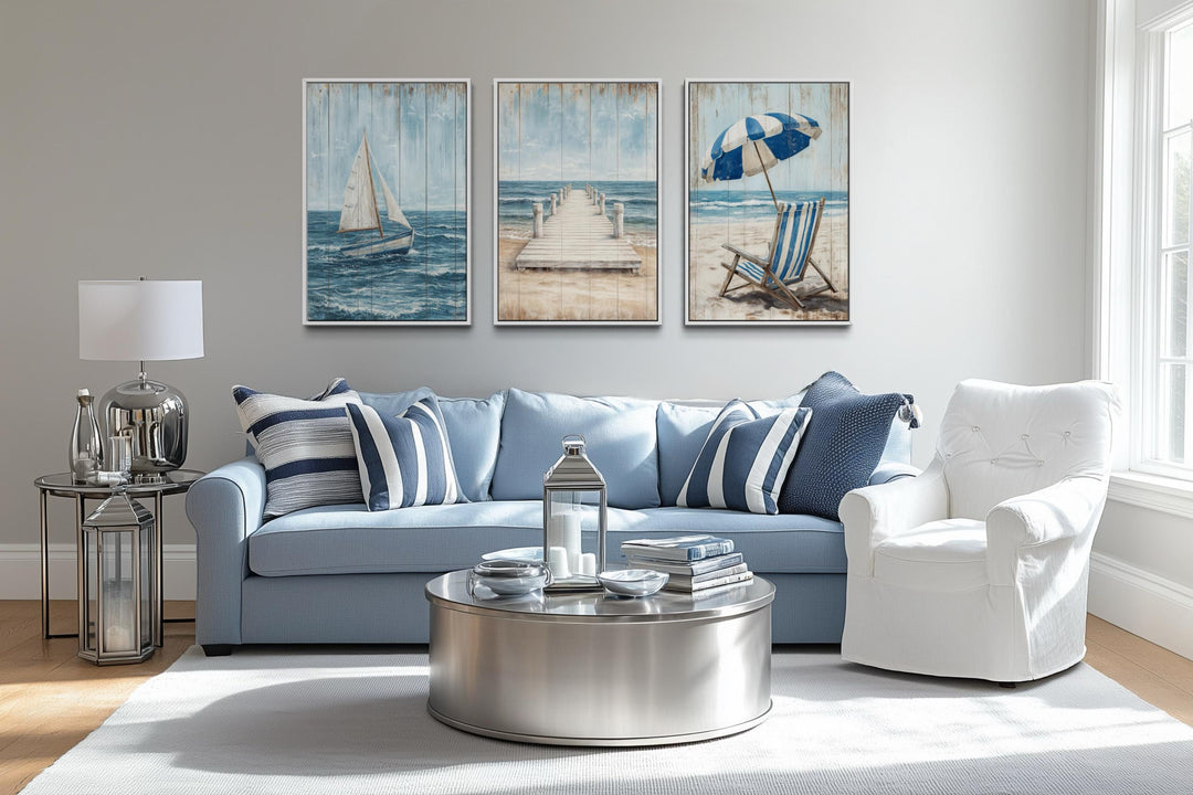 Set of three coastal canvas prints featuring a sailboat, pier, and beach umbrella over a blue sofa.