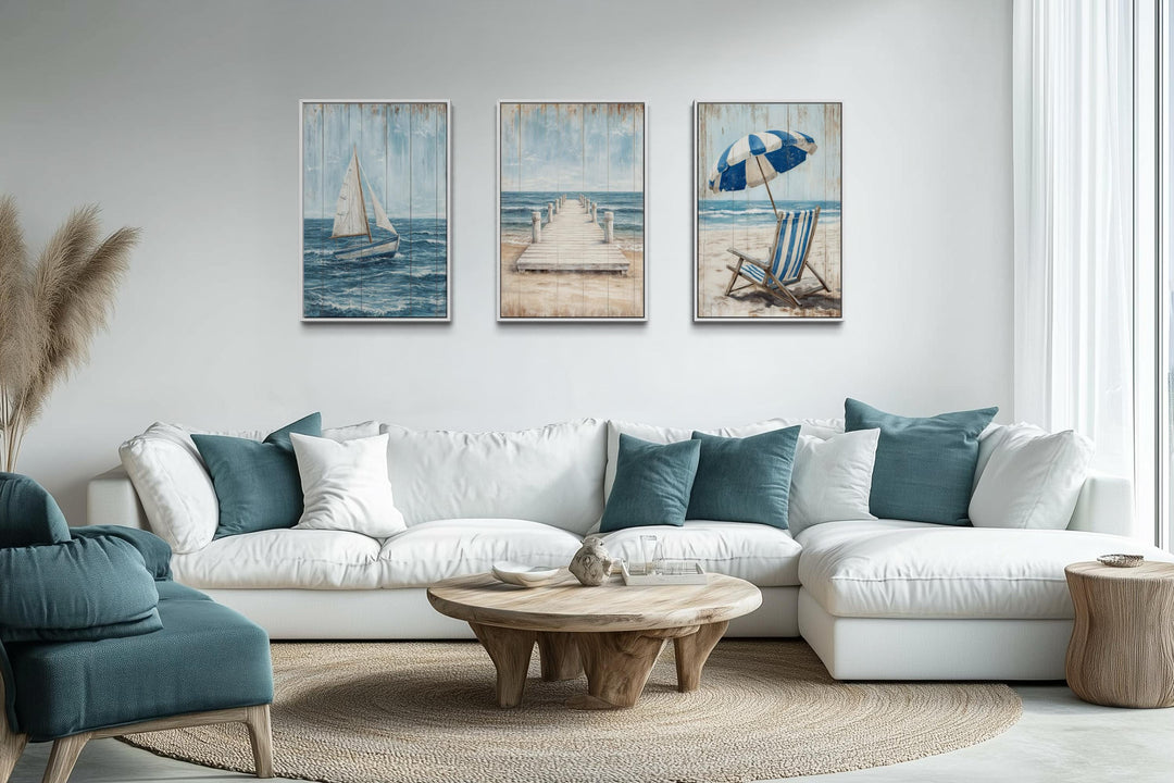 Three vertical nautical canvas prints featuring a sailboat, pier, and beach chair above a white sofa.