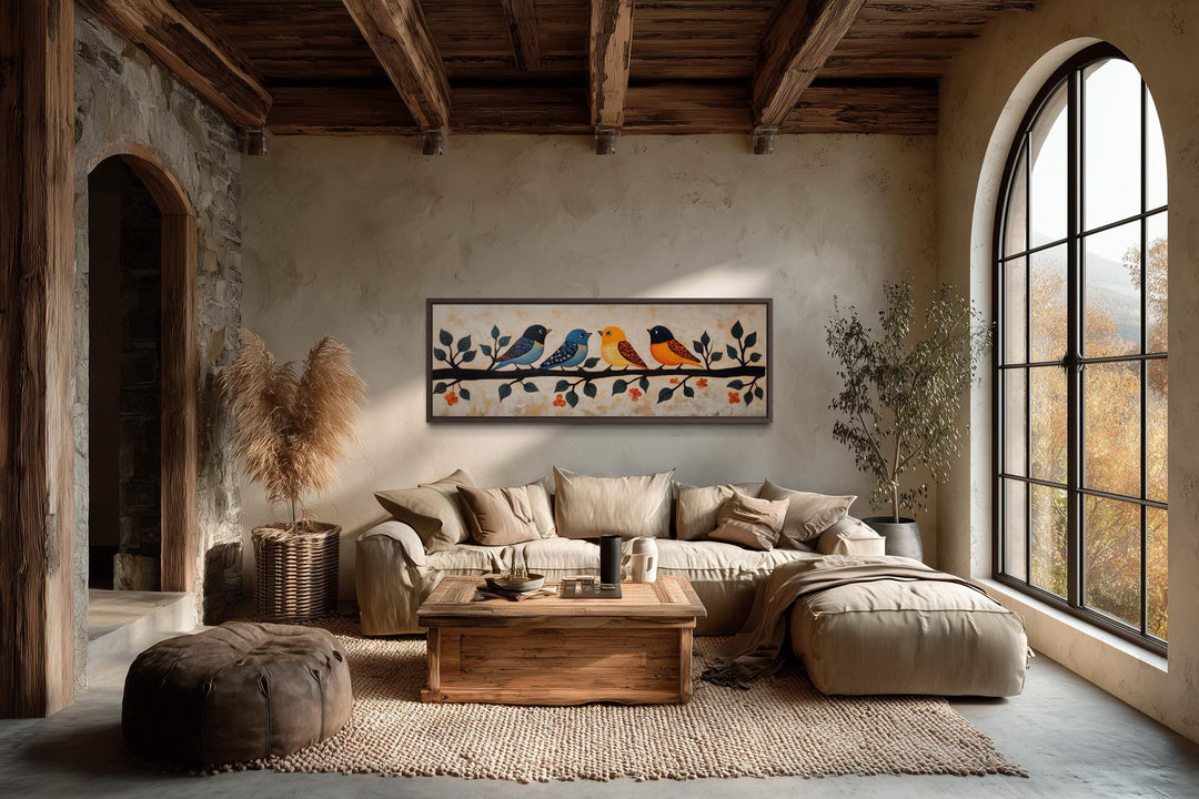Long narrow folk style canvas print of colorful birds on a branch above a neutral sofa in a room.