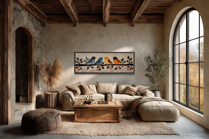 Long narrow folk style canvas print of colorful birds on a branch above a neutral sofa in a room.