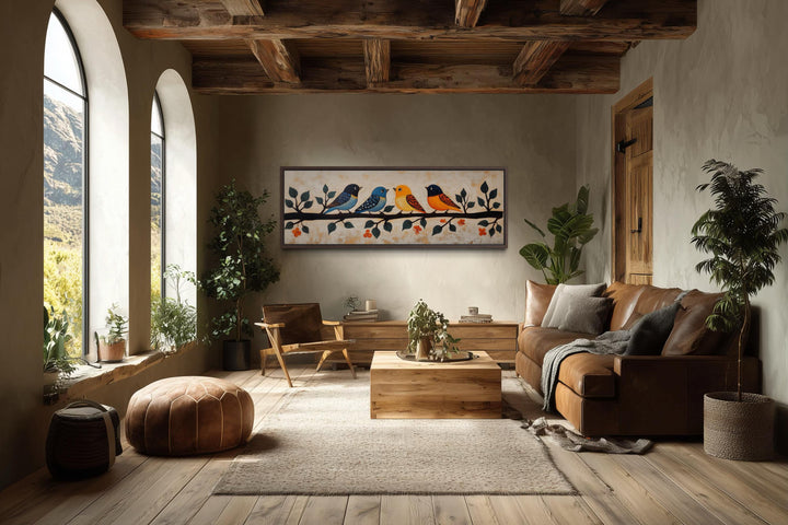Long narrow folk art canvas print of colorful birds on a branch above a sofa in a rustic room.