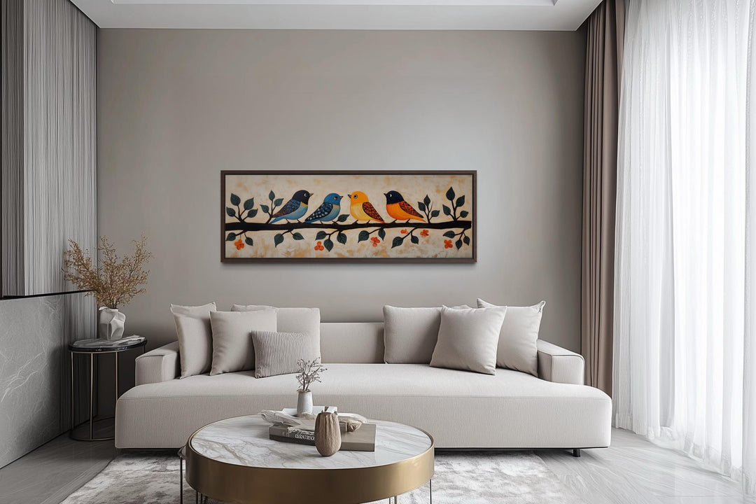 Long narrow folk style canvas print of four colorful birds on a branch above a modern beige sofa.