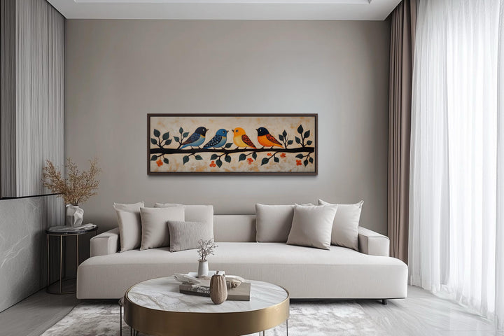Long narrow folk style canvas print of four colorful birds on a branch above a modern beige sofa.