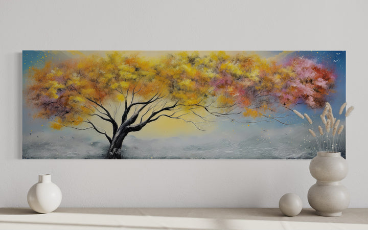 Horizontal canvas wall art of a vibrant tree with yellow, orange, and pink leaves on a white wall.