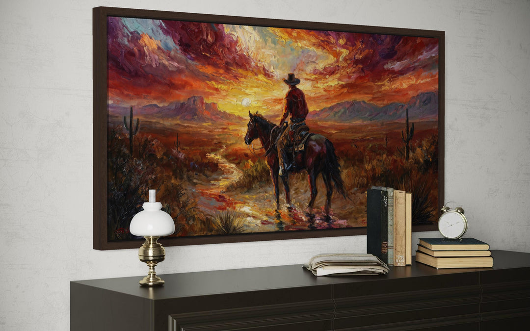 A framed canvas painting of a cowboy on horseback riding through a desert at sunset.