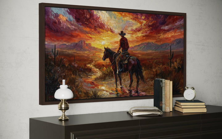A framed canvas painting of a cowboy on horseback riding through a desert at sunset.