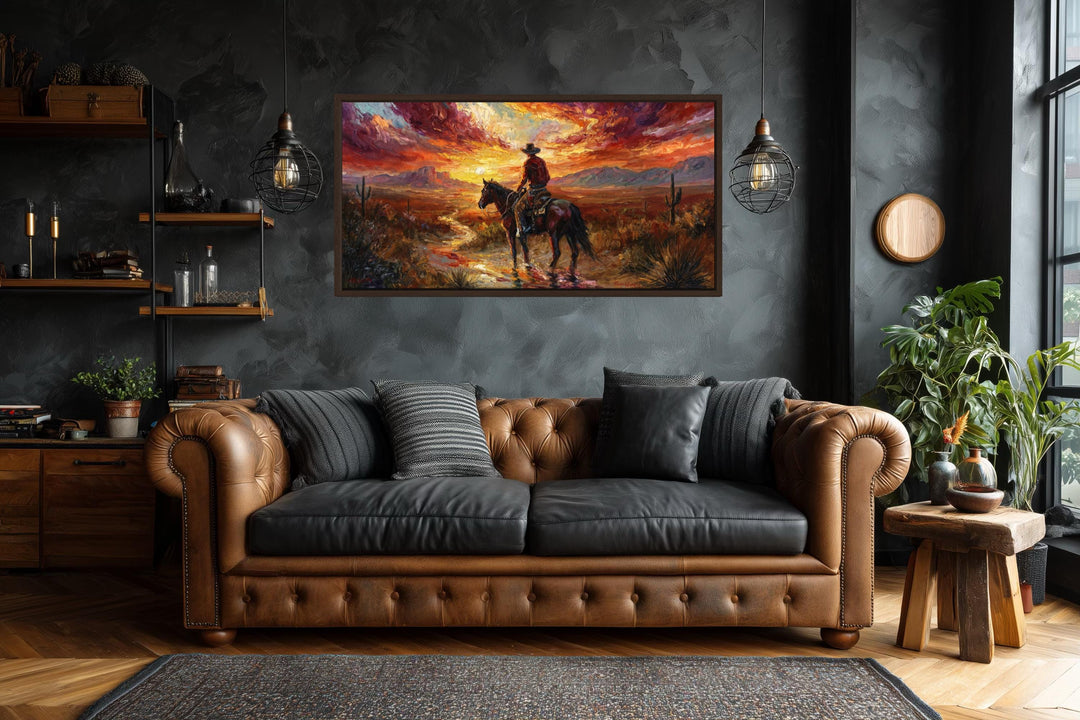 A framed canvas of a lone cowboy riding through a desert at sunset above a brown leather sofa.