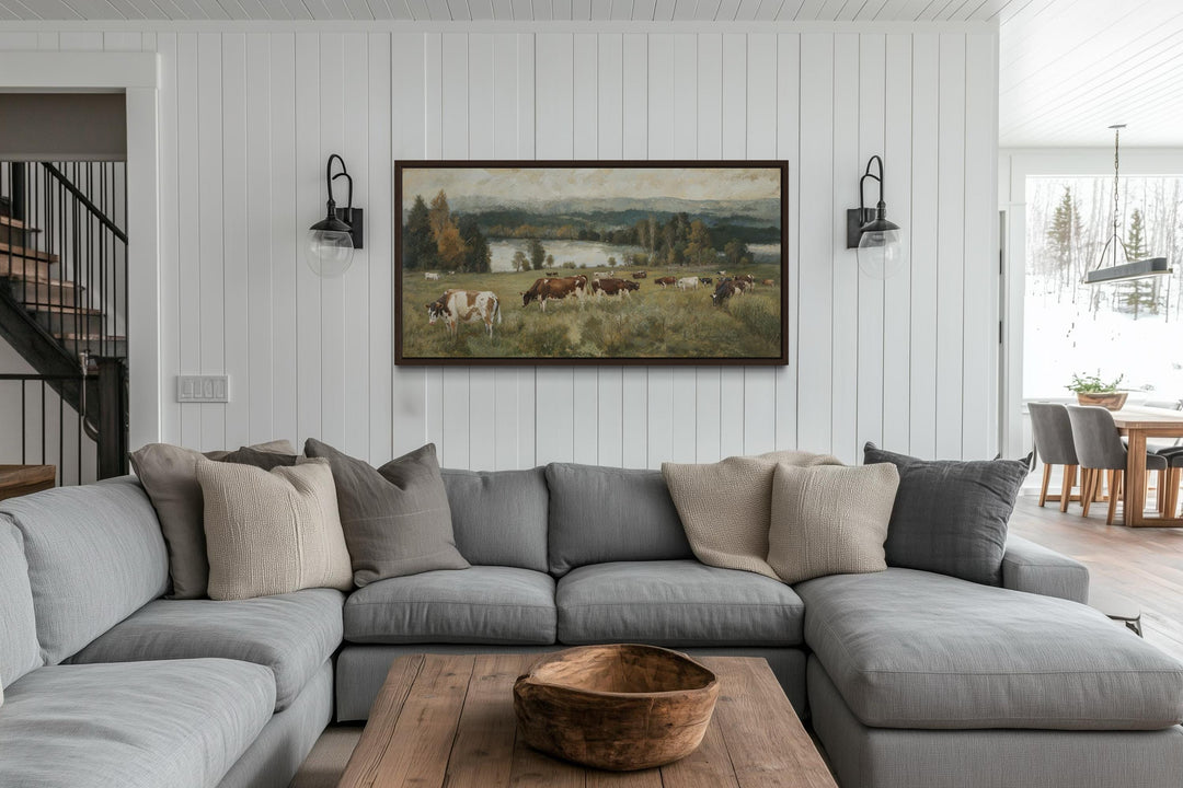 A large horizontal canvas of cows grazing in a pasture, framed above a modern sectional sofa.
