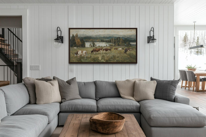 A large horizontal canvas of cows grazing in a pasture, framed above a modern sectional sofa.
