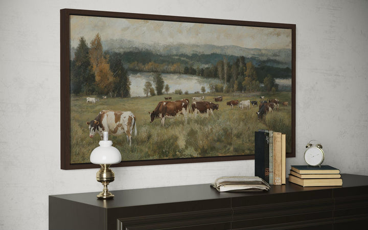 A framed horizontal canvas painting of cows grazing in a lush pasture by a lake.