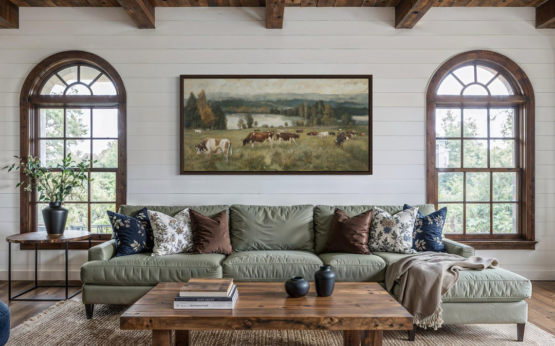A large horizontal canvas painting of cows grazing in a lush pasture above a green sofa.