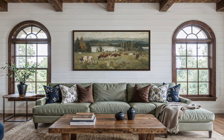 A large horizontal canvas painting of cows grazing in a lush pasture above a green sofa.