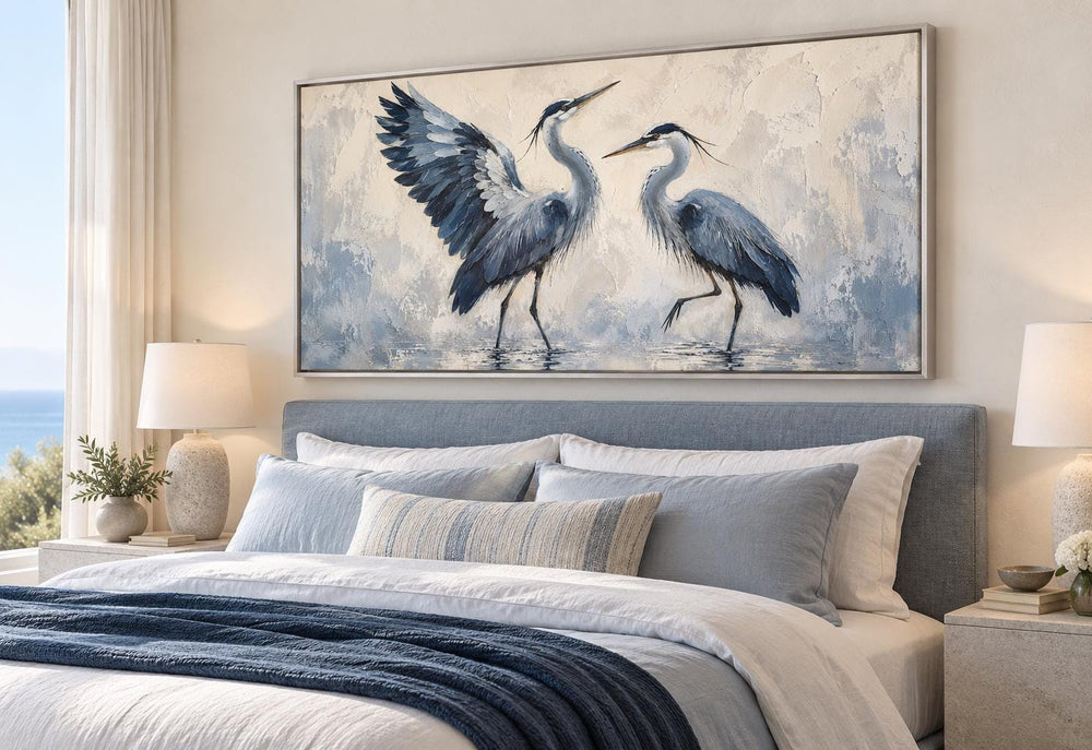 Dancing Blue Herons canvas wall art above a bed with blue and white bedding and bedside lamps.