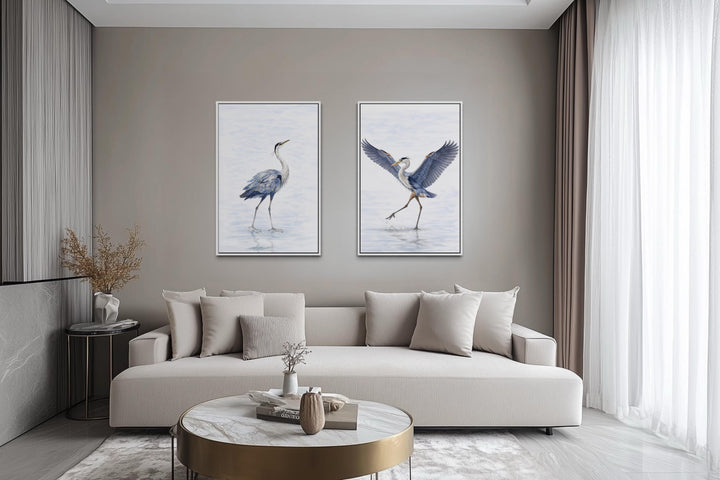 Set of two watercolor heron bird canvas prints hanging above a neutral sofa in a modern living room.