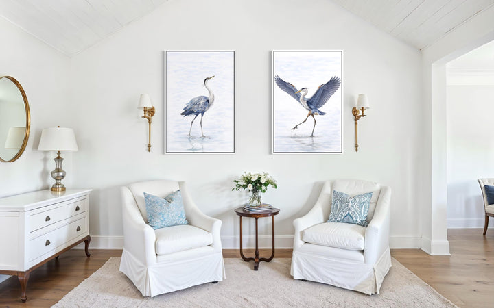 Set of two watercolor heron canvas prints in white frames hanging above white armchairs.