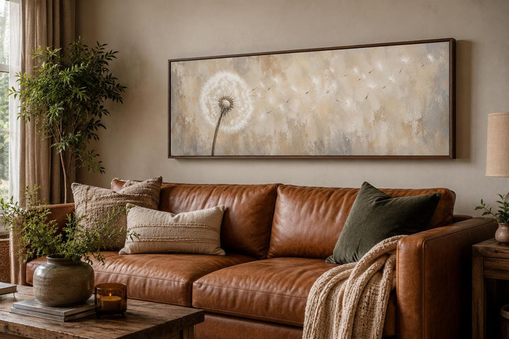 Long horizontal neutral canvas art of dandelion seeds blowing in the wind above a leather sofa.