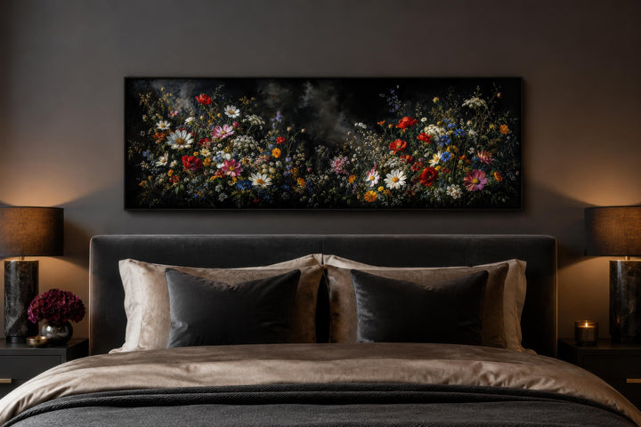 Long narrow canvas print of colorful wildflowers on a dark background above a bed in a moody room.