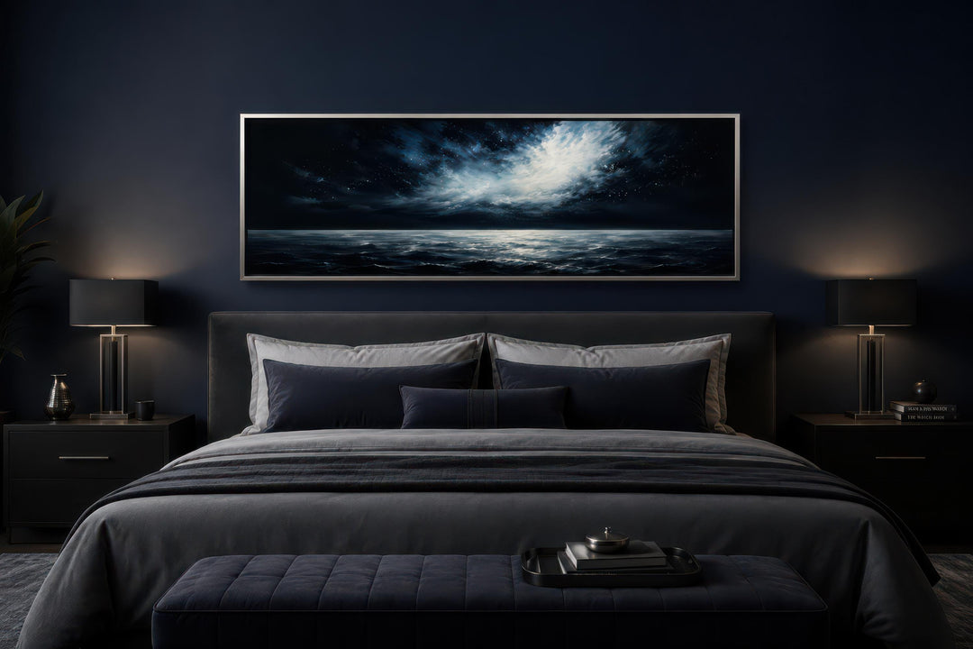 Dark navy moonlit ocean seascape painting in a silver frame above a bed in a moody bedroom.