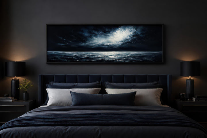 Dark navy moonlit ocean seascape painting in a black frame above a bed in a moody bedroom.