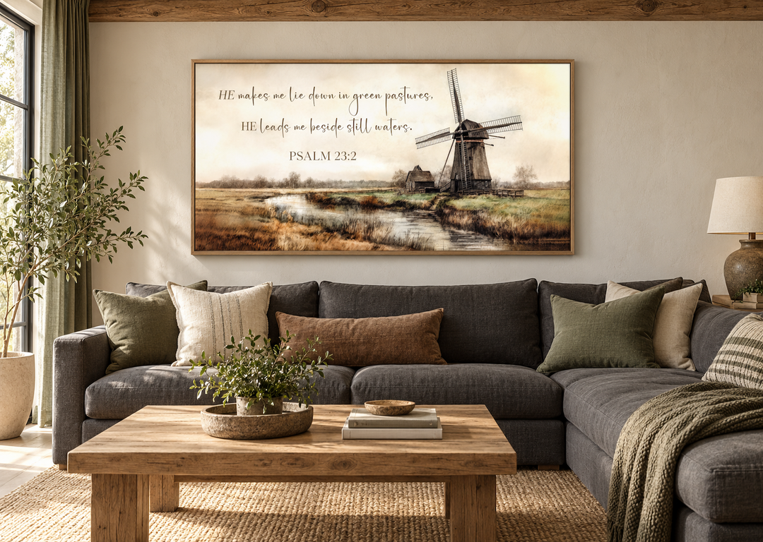 Psalm 23:2 scripture wall art featuring a windmill and river landscape above a gray sectional sofa.