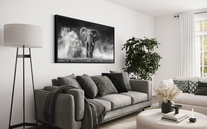 A framed black and white canvas print of an elephant in the dust hangs above a modern gray sofa.
