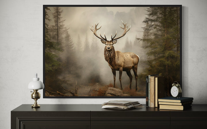 Framed elk painting in a misty fir forest, displayed on a dark dresser with books and a lamp.