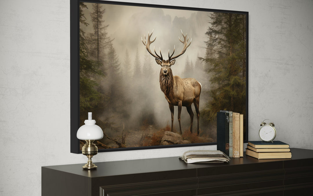 Framed elk painting in a misty fir forest on a dark wood dresser with books and a clock.