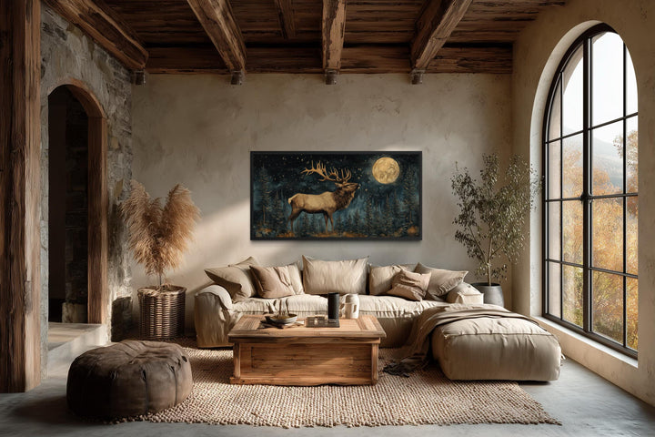 A horizontal canvas print of an elk in a moonlit forest hangs in a rustic, earth-toned living room.