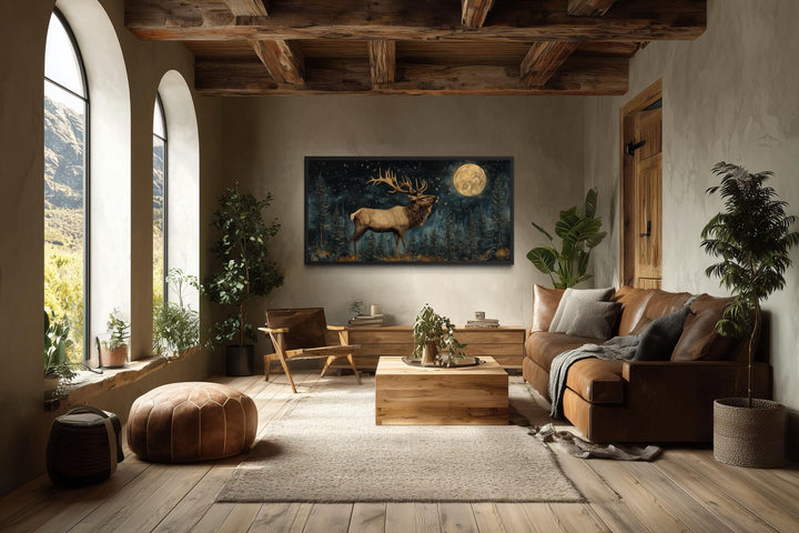 A horizontal canvas of an elk in a moonlit forest hangs in a rustic living room with wood beams.