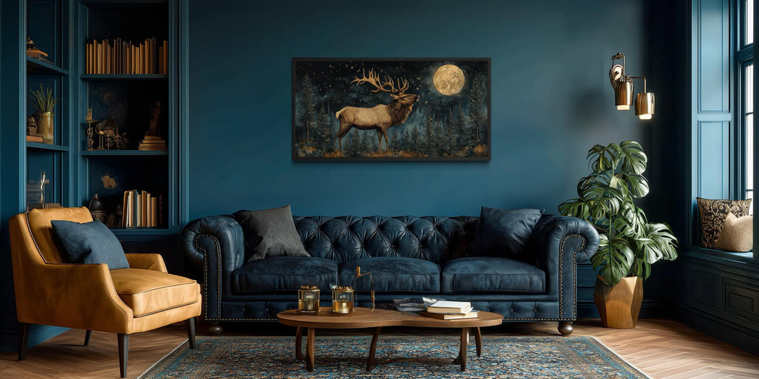 Horizontal wall art of an elk in a night forest with a full moon above a blue tufted sofa.
