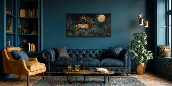 Horizontal wall art of an elk in a night forest with a full moon above a blue tufted sofa.